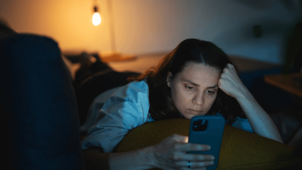 Mujer en la oscuridad de su habitación mirando fijamente la pantalla iluminada de su teléfono móvil con expresión de preocupación y cansancio.