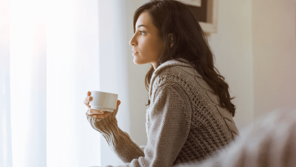 Mujer joven sosteniendo una taza de café junto a una ventana con luz natural, en un momento de pausa y desconexión digital.