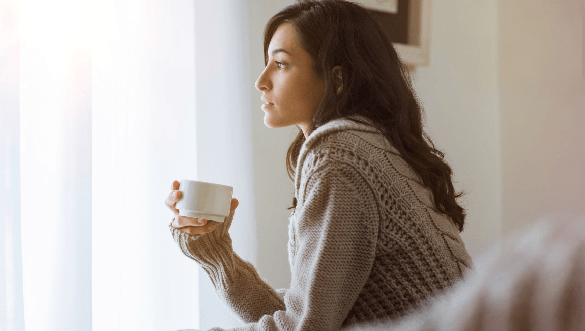 Mujer joven sosteniendo una taza de café junto a una ventana con luz natural, en un momento de pausa y desconexión digital.