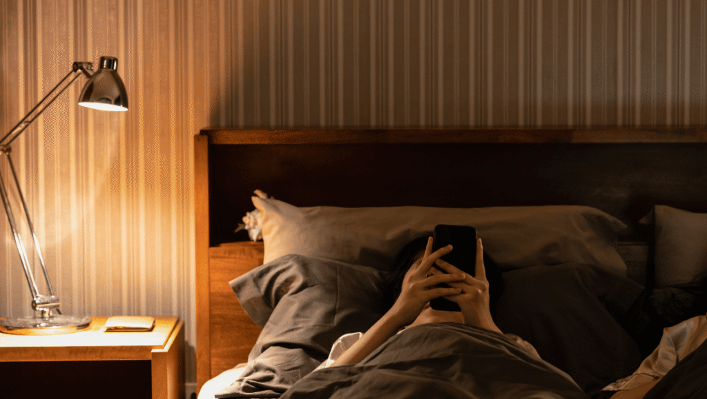 Mujer acostada en la cama de noche mirando el celular, cansada por la exigencia digital